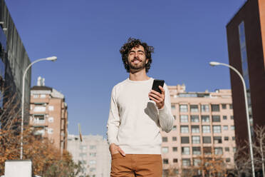 Smiling man with eyes closed holding smart phone under blue sky - EBBF07876