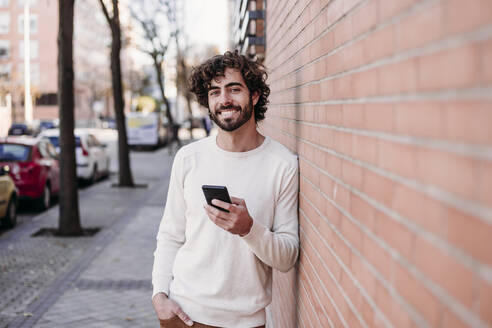 Lächelnder junger Mann mit Smartphone an der Wand lehnend - EBBF07877