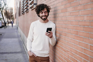 Glücklicher junger Mann mit Smartphone an der Wand lehnend - EBBF07878