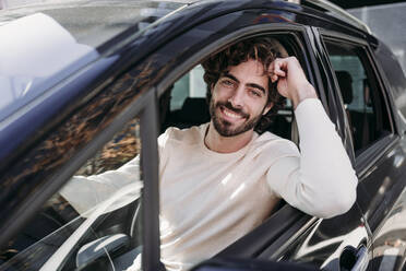 Smiling young man with beard leaning on car window - EBBF07891