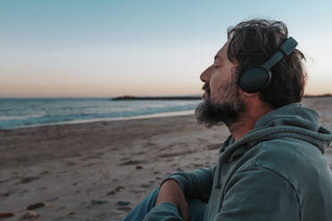 Älterer Mann mit Kopfhörern, der am Strand sitzt und Musik hört - SIPF02889