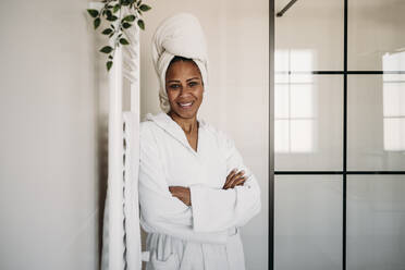 Happy mature woman with arms crossed standing in bathroom - EBBF07898