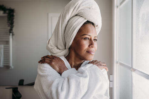 Mature woman wearing bathrobe looking through bathroom window - EBBF07909