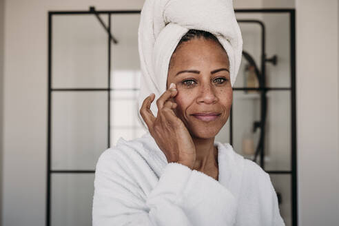 Smiling woman applying moisturizer on face in bathroom at home - EBBF07912