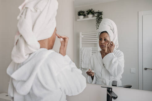 Mature woman applying moisturizer on face in bathroom at home - EBBF07914