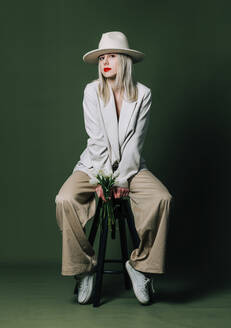 Woman with hat sitting on stool in front of green background - VSNF00409