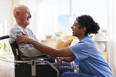 Fröhliche Physiotherapeutin im Gespräch mit einem älteren Mann, der im Rollstuhl sitzt - EBSF02683