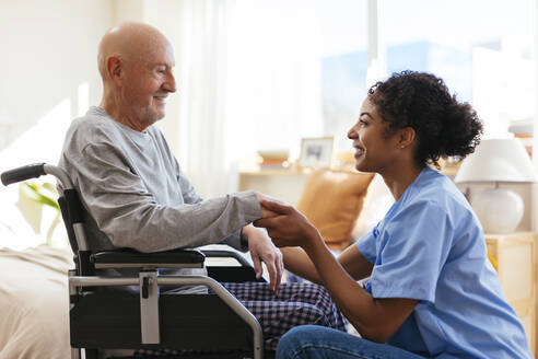 Glückliche Physiotherapeutin hält die Hand eines älteren Mannes, der im Rollstuhl sitzt - EBSF02684