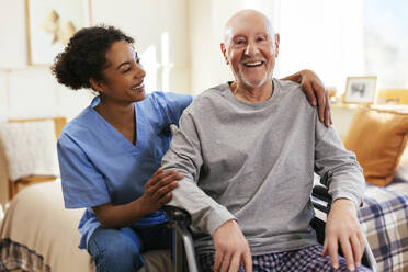 Happy senior man sitting on wheelchair by nurse at home - EBSF02685