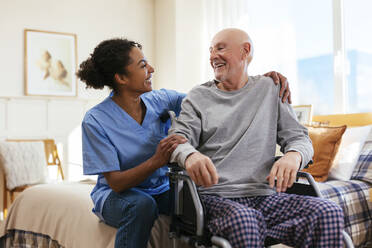 Happy nurse with senior man sitting on wheelchair in bedroom at home - EBSF02686