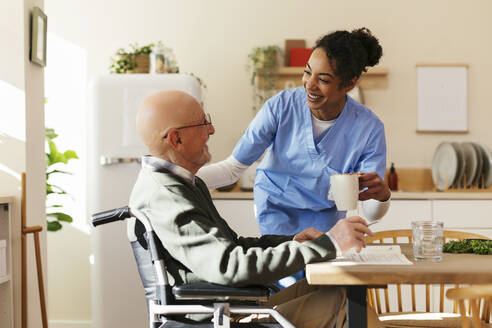 Smiling caretaker giving mug to senior man on wheelchair at home - EBSF02696