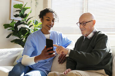 Smiling senior man sharing smart phone with caretaker at home - EBSF02709