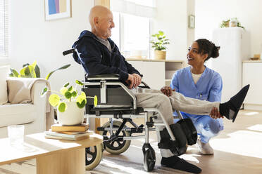 Smiling physiotherapist examining senior man's leg on wheelchair at home - EBSF02716