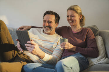 Happy mature couple watching tablet PC sitting on sofa at home - JOSEF16645