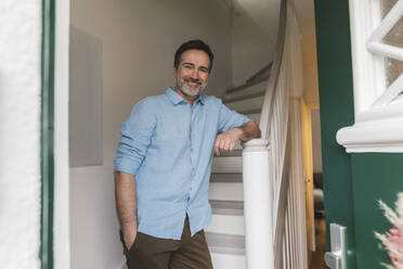 Lächelnder reifer Mann mit Hand in der Tasche steht in der Nähe der Treppe zu Hause - JOSEF16669