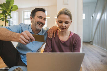 Happy husband holding credit card by woman using laptop at home - JOSEF16746