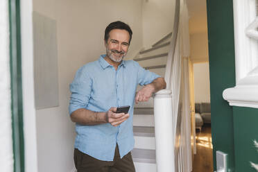 Lächelnder reifer Mann mit Smartphone vor einer Treppe zu Hause stehend - JOSEF16811