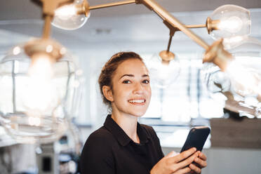 Lächelnde junge Geschäftsfrau mit Smartphone unter beleuchteten Lichtern - JOSEF16844