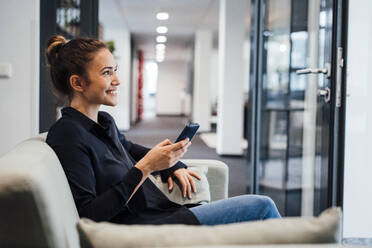 Lächelnde junge Geschäftsfrau sitzt mit Smartphone auf der Couch im Büro - JOSEF16857
