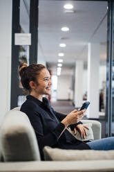 Happy businesswoman sitting with mobile phone in office - JOSEF16858