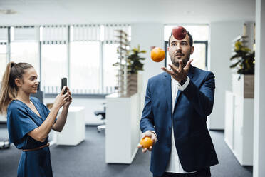 Businesswoman taking photo using smart phone of colleague juggling fruits in office - JOSEF16859