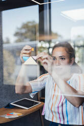 Young businesswoman sticking adhesive notes on glass - JOSEF16875