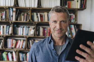 Smiling man with tablet PC standing in front of bookshelf - KNSF09643