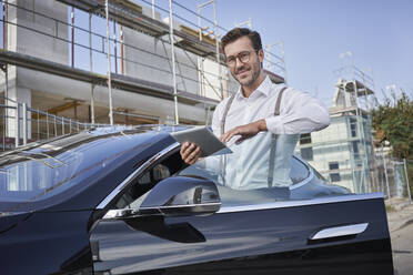 Lächelnder Architekt mit Tablet-PC neben einem Auto auf einer Baustelle - RORF03337