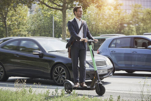 Smiling businessman standing on electric push scooter on road - RORF03344