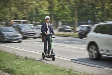 Junger Geschäftsmann mit Helm, der auf einem Elektroroller auf der Straße fährt - RORF03346