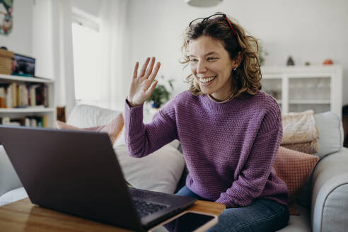 Happy woman waving on video call through laptop at home - DMGF01020