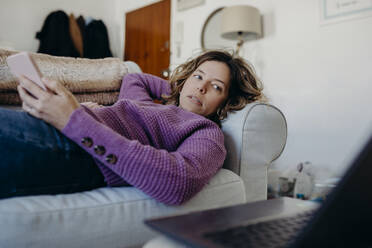 Woman holding smart phone lying on sofa at home - DMGF01023