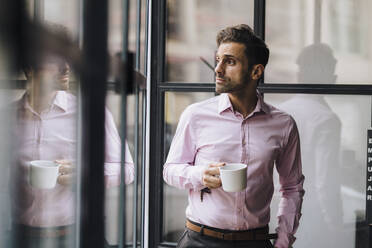 Thoughtful businessman holding coffee cup in front of glass wall - JOSEF16892