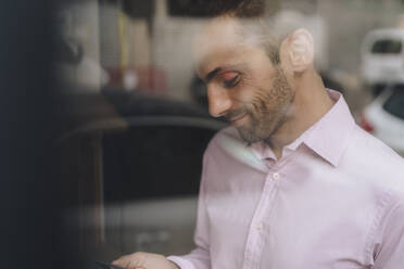 Smiling mature businessman using smart phone seen through glass - JOSEF16899