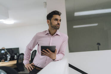 Thoughtful businessman with tablet PC leaning on wall in office - JOSEF16923