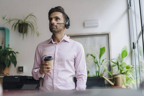 Nachdenklicher reifer Geschäftsmann mit Headset und Einweg-Kaffeetasse im Büro - JOSEF16929