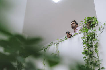 Nachdenklicher reifer Geschäftsmann mit Mobiltelefon, der sich an eine Wand mit Pflanzen lehnt - JOSEF16937