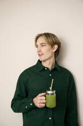 Young man holding green smoothie against beige background - RSKF00032