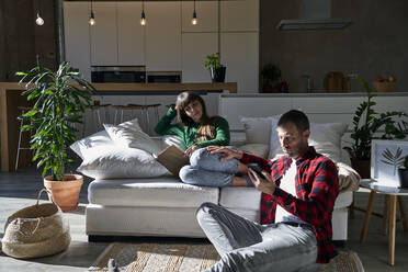 Mann benutzt Smartphone mit Freundin, die zu Hause auf der Couch ein Buch liest - VEGF06202