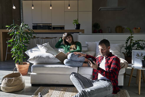 Man using smart phone with girlfriend reading book on couch at home - VEGF06202