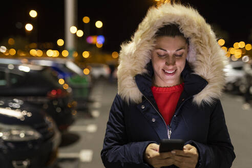 Happy woman wearing parka hood using smart phone at night - LJF02477