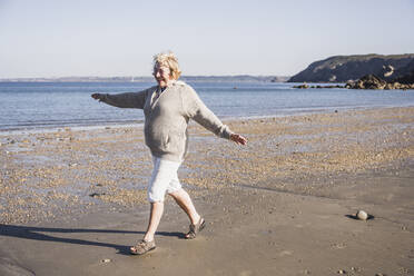 Sorglose ältere Frau mit ausgestreckten Armen, die an einem sonnigen Tag am Strand spazieren geht - UUF28154