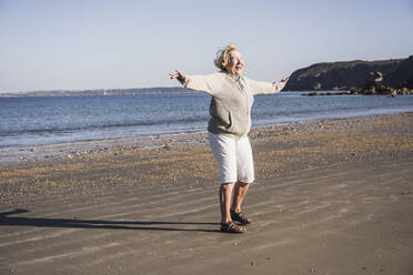 Fröhliche ältere Frau mit ausgestreckten Armen am Strand im Urlaub - UUF28157