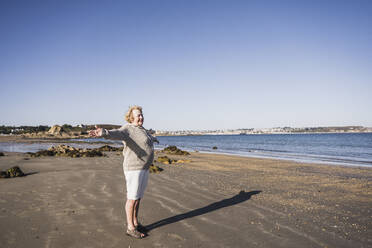 Sorglose ältere Frau mit ausgestreckten Armen am Strand stehend - UUF28161