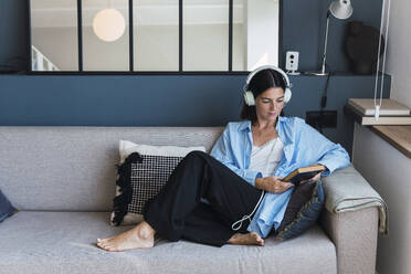 Junge Frau mit Buch, die zu Hause über Kopfhörer Musik hört - PNAF04971