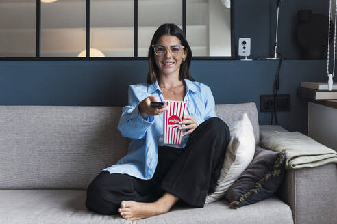 Lächelnde Frau mit Popcorn und TV-Fernbedienung auf dem Sofa zu Hause - PNAF04981