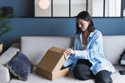 Young woman opening package on sofa at home - PNAF04994