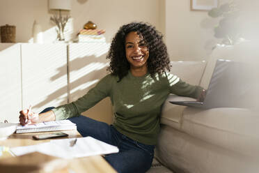 Glückliche Freiberuflerin mit lockigem Haar sitzt auf der Couch im Home Office - EBSF02727