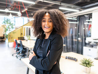 Positive afroamerikanische Frau mit Afrofrisur in formellem Outfit, die lächelt und in die Kamera schaut, während sie mit verschränkten Armen im Büro steht - ADSF43095