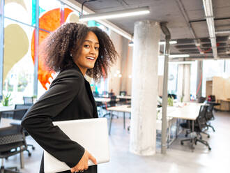 Positive afroamerikanische Frau mit Afrofrisur in formeller Kleidung, stehend in einem modernen Arbeitsbereich mit Netbook - ADSF43096